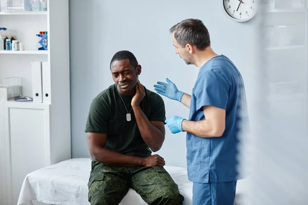 年轻的黑人士兵坐在现代军医院的医疗沙发上 告诉医生颈部疼痛 — 图库照片