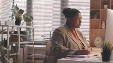 Yan görünüm, not defterine not alan ve ofis masasında otururken kablosuz klavyeye yazı yazan Afro-Amerikan genç ve büyük boy bir kadının yavaş belini kaldırıyor.
