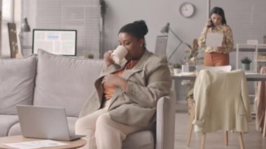 Modern ofisteki kanepede oturan müşteri ya da iş ortağıyla sohbet eden orta yaşlı, iri yarı bir zenci kadın videosu. Sekreteri arka planda bulanık telefon görüşmeleri yapıyor.