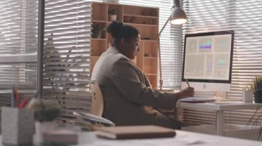 İş yerinde bilgisayarın başında oturan ve not defterine not alan siyahi kadının orta boy bir yan görünüm yavaşlığı