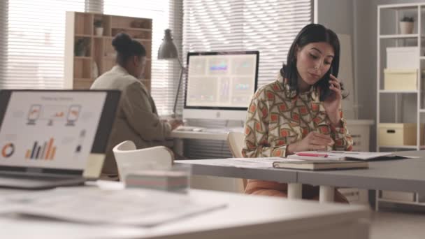 慢吞吞地等着年轻的中东女商人一边看文件 一边用手机与客户交谈 她的非裔美国秘书背景在电脑上工作 — 图库视频影像