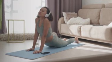 Kulaklıklı, evde yoga yaparken yukarı doğru köpek pozu veren yavaş, melez kız.
