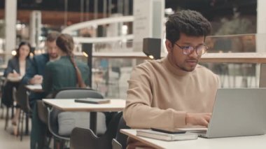 Zekice giyinen ve gözlüklü, yemek masasında dizüstü bilgisayarda çalışan iki ırklı genç adamın belini kaldır.