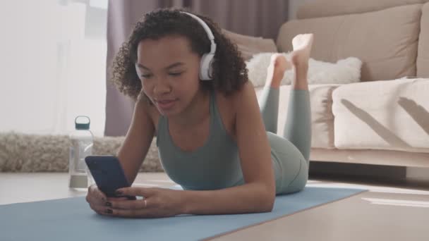 身着运动服 头戴耳机的美女慢吞吞地躺在家里的垫子上 在智能手机上打滚 听音乐 在运动间隙休息 — 图库视频影像