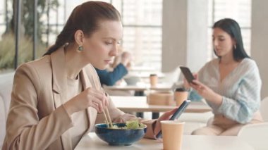 Yemek salonunda panoramik pencereli yemek çubuklarıyla yemek yerken akıllı telefon kullanan beyaz bir kadının belini kaldır.