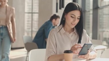 Kafede panoramik pencereli bir masada tek başına otururken akıllı telefon kullanan çekici, genç, esmer bir kadının belini kaldır.