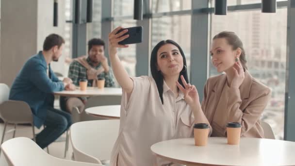 2人の若い美しい多民族女性の親友の中低速パノラマの窓とフードコートでテイクアウトコーヒーを飲みながらスマートフォンで自撮り写真を撮る — ストック動画