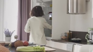 Çift ırklı genç bir kadının yiyecek ürünlerini buzdolabına koyarken orta derecede yavaşlaması, yiyecek alışverişinden eve yeni geldi.