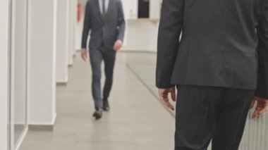 Orta kısımda, takım elbise ve kravat takmış iki tanınmayan iş adamının modern iş merkezinin koridorunda yürürken el sıkışarak birbirlerini selamlamalarını izliyoruz.