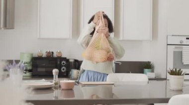 Gıda alışverişinden eve gelip, taze meyve ve sebzeleri net poşetinden çıkartan melez genç bir kadın.