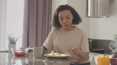 Orta yavaş kıvırcık saçlı, kıvırcık saçlı, evde kahvaltı yaparken arkadaşlarıyla mesajlaşan çift ırklı bir kadın.