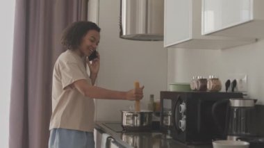 Orta yavaş gülümseyen, iç çamaşırlı, çift cinsiyetli bir kadının mutfakta spagetti pişirirken akıllı telefonuyla sohbet etmesi.