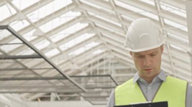 Düşük açılı göğüs yukarı, inşaat alanı boyunca inşaat planıyla yürüyen sert şapka ve yansıtıcı yelek giyen ustabaşının yavaş yavaş yukarı doğru, check-up yapıyor.