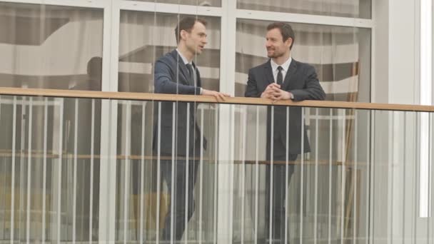 現代的なビジネスセンターでの会話を持つガラスの手すりに立ってエレガントなFormalwearで2人の白人ビジネスマンの完全な長さの低速 — ストック動画