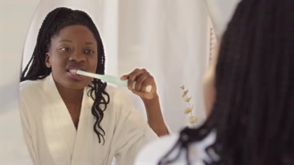 身穿白色浴衣的年轻非洲裔美国妇女从肩部向上慢慢地看着镜子里自己的倒影 同时彻底地刷牙 — 图库视频影像