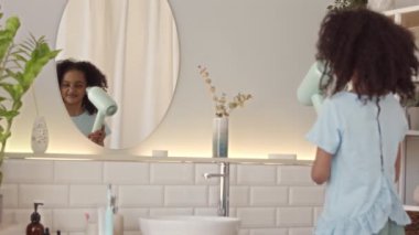 Arkadan bakıldığında, 6 yaşında, şık, minimalist bir banyoda aynanın önünde duran kıvırcık saçlarını kurutan güzel, çift cinsiyetli bir kızın orta derece yavaşlığı.