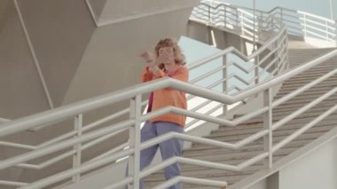 Renkli y2k kıyafetli beyaz bir kız merdivenlerde serbest dans ediyor.
