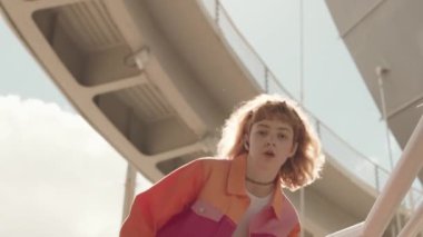 Alçak açılı, beyaz tenli, pembe bloklu, y2k ceketli bir kız. Açık havada güneş ışığında dans ediyor. Kameraya bakıyor.