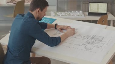 Ofiste ev planı çizerken cetvel ve kalem kullanan, konsantre beyaz erkek mimarın dikiz görüşü orta yavaştır.