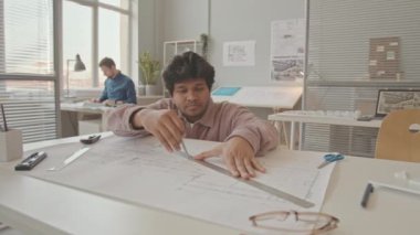 Ofis masasının yanında oturan profesyonel erkek çift ırklı mimarın yavaş çekimleri ile orta ölçekli bir yakın çekim.