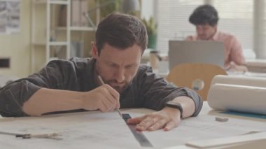 Modern ofiste inşa planlarını çizerken kalem ve cetvel kullanan konsantre erkek mimarın yavaş çekimleri.