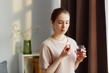 Vitamin kapsülü tutan ve evinde bir bardak suyla takviye alan genç bir kadının minimal portresi.