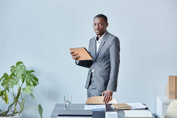 Minimal portrait of professional black executive looking at camera while standing against blue wall in office wearing elegant suit, copy space
