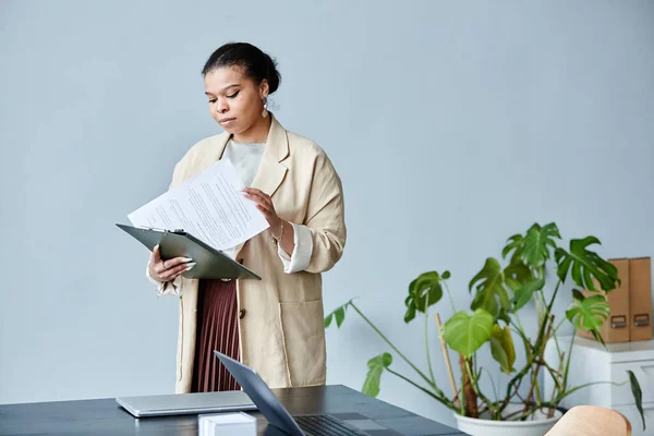 最低限の灰色の背景に対してオフィスに立っている間 文書を読んでエレガントな黒のビジネスマンの女性のウエストアップ肖像画 コピースペース — ストック写真