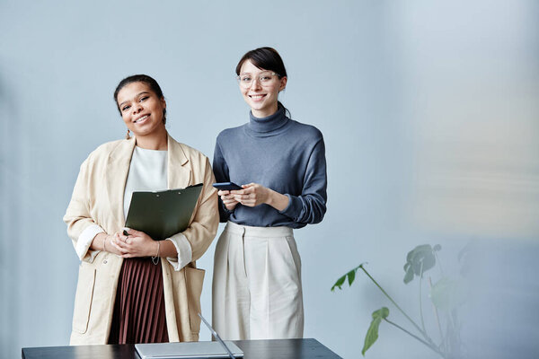 Minimal portrait of two young businesswomen smiling at camera while standing in office against simple grey background, copy space