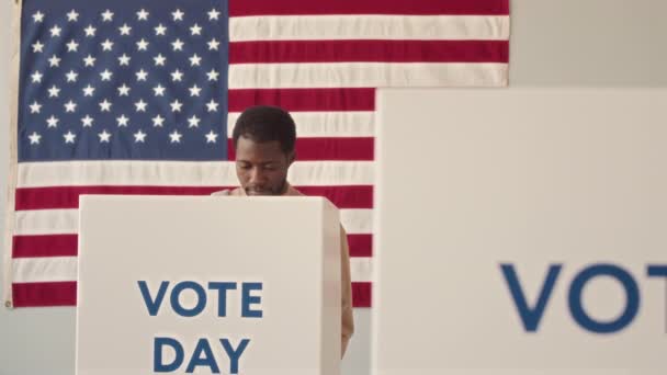 選挙区で米国の旗に反対する投票ブースに来て 彼の投票を内側に置く若いアフリカ系アメリカ人の男性の中低速 — ストック動画