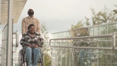 Ağır ağır, iri yarı, özürlü Afro-Amerikan kadın ve tekerlekli sandalyesini taşıyan erkek arkadaşı dışarıda yürüyüp sohbet ediyorlar.