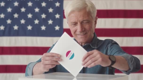 照片中快乐的白人老人把选票放进投票箱 面对着站在投票站里高举美国国旗的摄像机微笑 — 图库视频影像