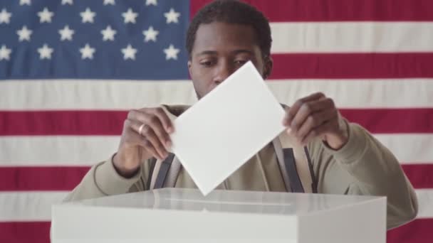 在投票站里 年轻的非裔美国人把选票放进投票箱 面对着高举美国国旗站在旁边的镜头笑着 把他的慢镜头放大 — 图库视频影像