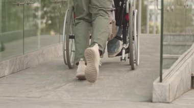 Arka manzara, tekerlekli sandalye kullanıcısının şehirde küçük bir yükselişe geçmesine yardım eden tanınmayan adamı yavaş yavaş yukarı kaldırır.