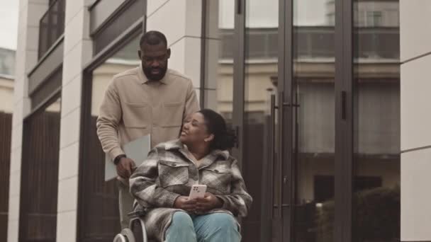 黒人男性の中低速彼の女性の同僚が車椅子に乗るのを助けるオフィスの外 — ストック動画