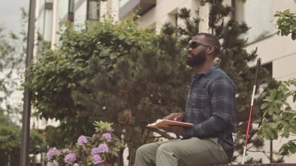 坐在公园长椅上阅读布莱叶盲文书籍的戴黑色眼镜的非洲裔美国青年盲人的慢动作 — 图库视频影像