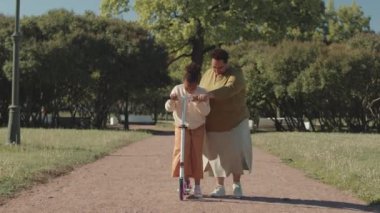 Ilık güneşli bir günde parkta 8 yaşındaki kızına scooter sürmesi için yardım eden Afrikalı Amerikalı kadın.