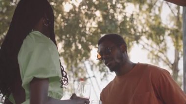 Düşük açılı orta yaşlı Afrikalı Amerikalı çift kırmızı şarap içiyor ve yazın ağaçların altında sohbet ediyorlar.