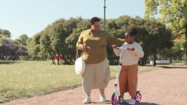 Büyük boy Afro-Amerikan kadın ve 8 yaşındaki kızı güneşli bir günde parkta yolda yürürler.