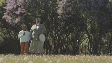 Kara hindiba tarlasında yürüyen, el ele tutuşan ve arka planda çiçek açan leylak ağaçlarıyla dondurma yiyen Afrikalı anne ve kız.