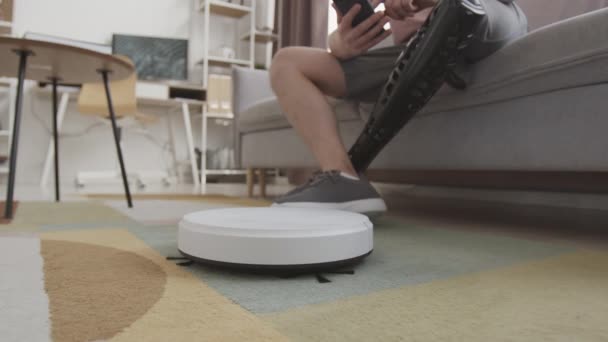 ロボット掃除機をスマートフォンに接続し リビングルームのソファに座っている義肢の脚を持つ認識できない男の作物の減速 — ストック動画