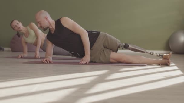白种人年轻男子假腿慢吞吞地在女教练的帮助下在垫子上做瑜伽 — 图库视频影像