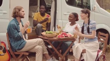 Bir grup neşeli çoklu etnik arkadaş yazın dışarıda piknik yapıyorlar karavanlarının yanında oturup sohbet ediyorlar ve bira içiyorlar.