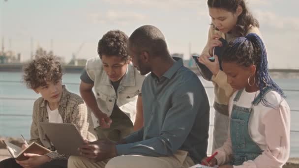 中等速度慢的非裔美国男教师和一群在课外上课 坐在阳光下的码头上 看着手提电脑屏幕的小学生 — 图库视频影像