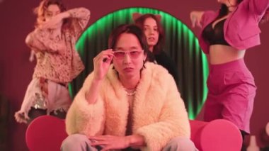 Moda pembe gözlüklü ve kürklü Asyalı hippi çocuk kameraya şatafatlı bir şekilde bakarken üç çekici kız retro stüdyosunda neon pembe ışıkla dans ediyor.