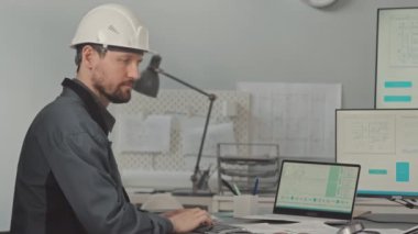 Beyaz şapkalı beyaz erkek tasarım mühendisinin ofis masasında bilgisayar ekranlarında teknoloji şemalarıyla otururken kameraya bakmasının orta yavaşlığı.