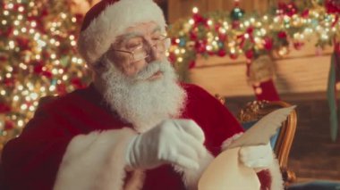 Medium slowmo of kind bearded Santa Claus in red costume sitting in comfortable velvet chair by fireplace reading kids letter
