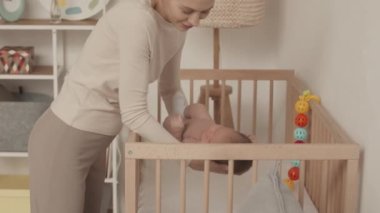 Medium slowmo of young Caucasian woman putting her beautiful newborn baby daughter in wooden crib to sleep