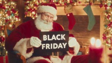 Medium slowmo portrait of smiling Santa in red costume sitting by decorated fireplace with Black Friday sign in hands