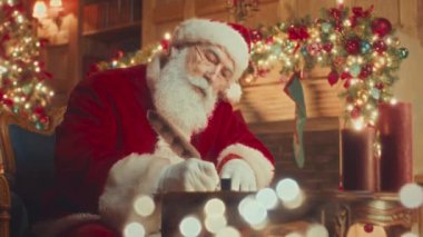 Medium slowmo of kind Santa in red costume using feather quill pen while writing letter to child sitting in cozy room with decorative shining lights